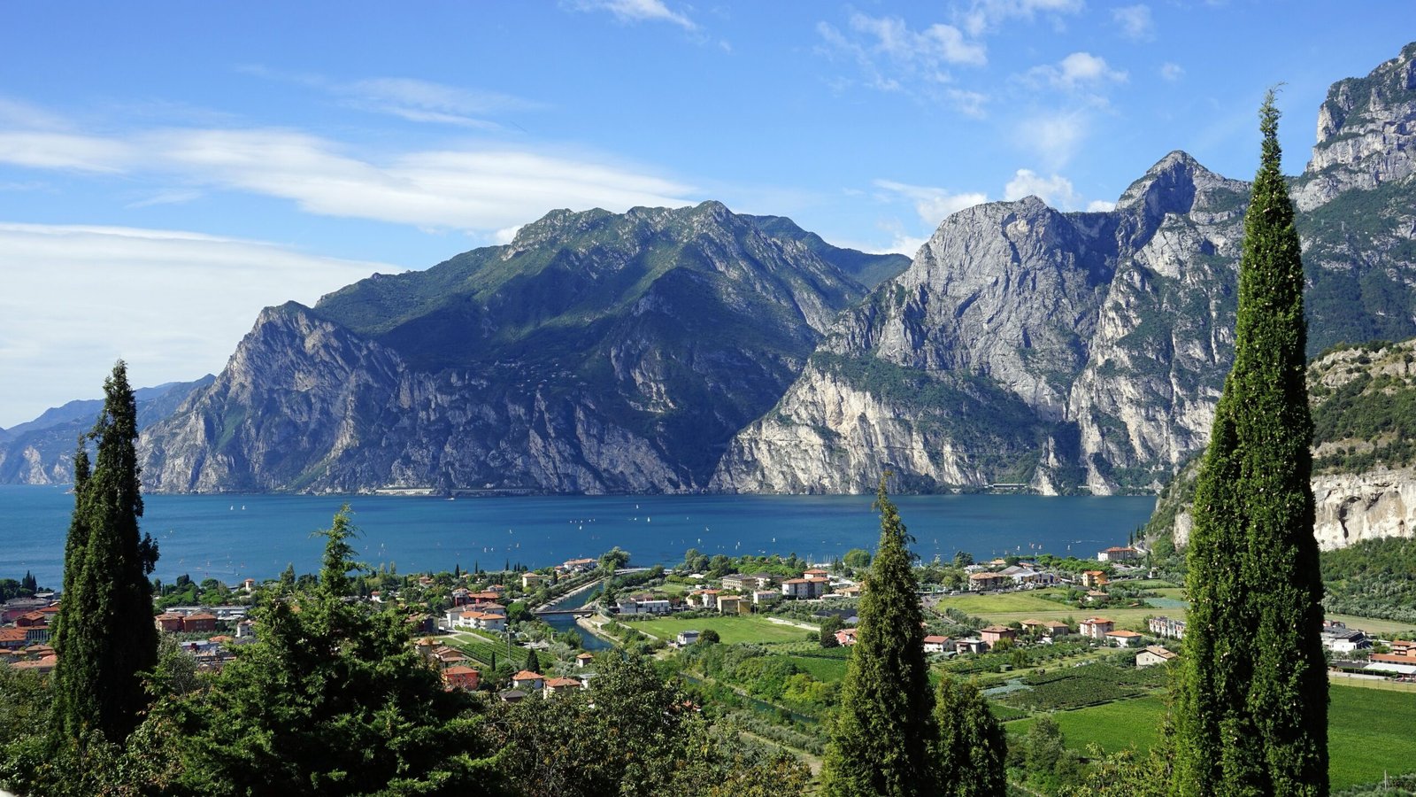 Northern Italy Scenic view of Lake Garda with surrounding mountains and lush green trees in the foreground.